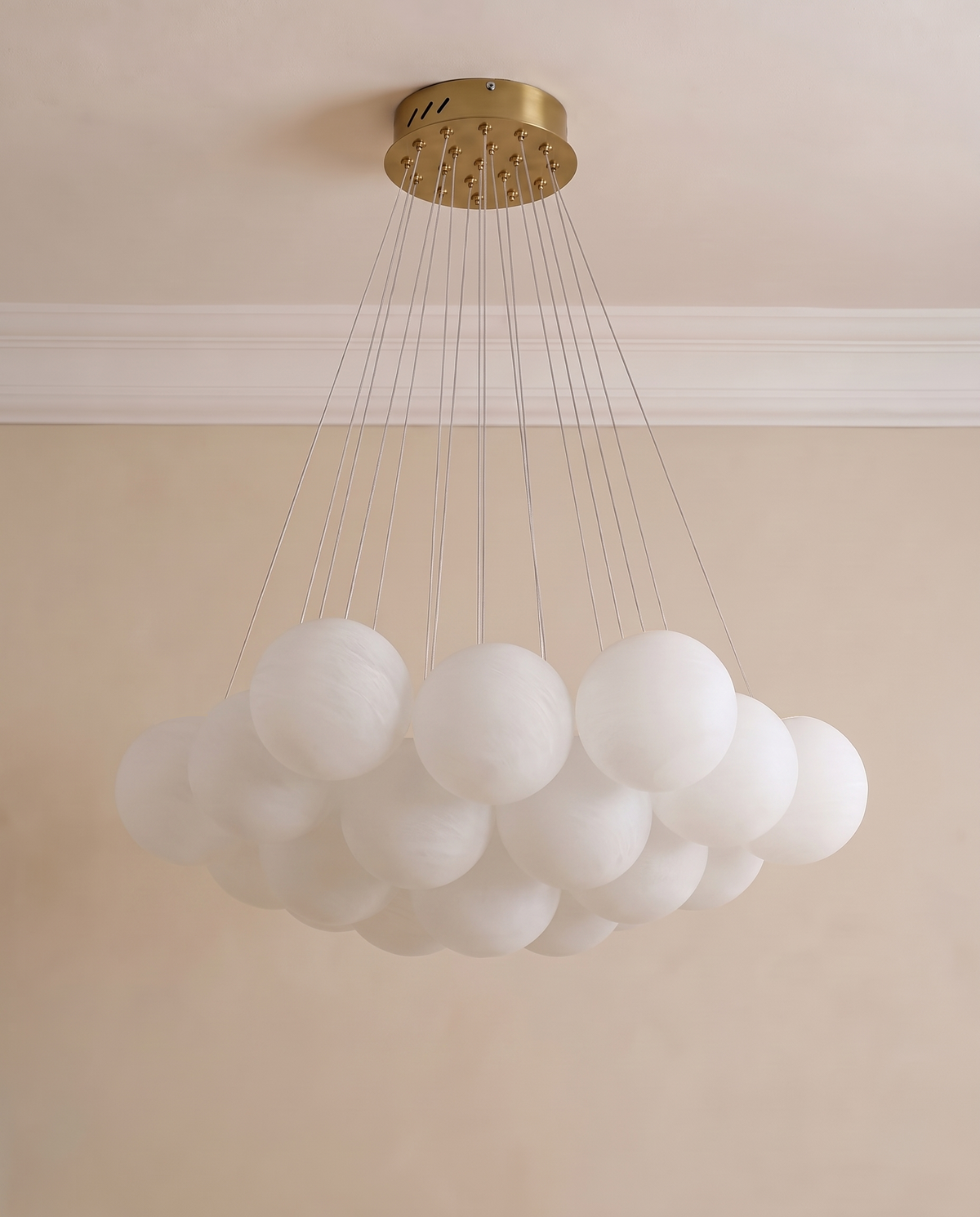 A modern cloud cluster chandelier with multiple frosted white acrylic globes suspended from a brushed gold circular ceiling canopy by thin cables, shown against a beige wall and ceiling.